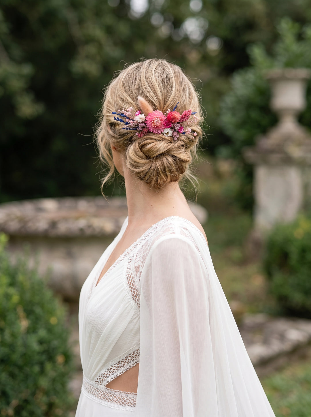 A woman in a white wedding dress shown from the back in an outdoor garden setting, wearing a floral hair accessory with pink, purple, and green flowers.