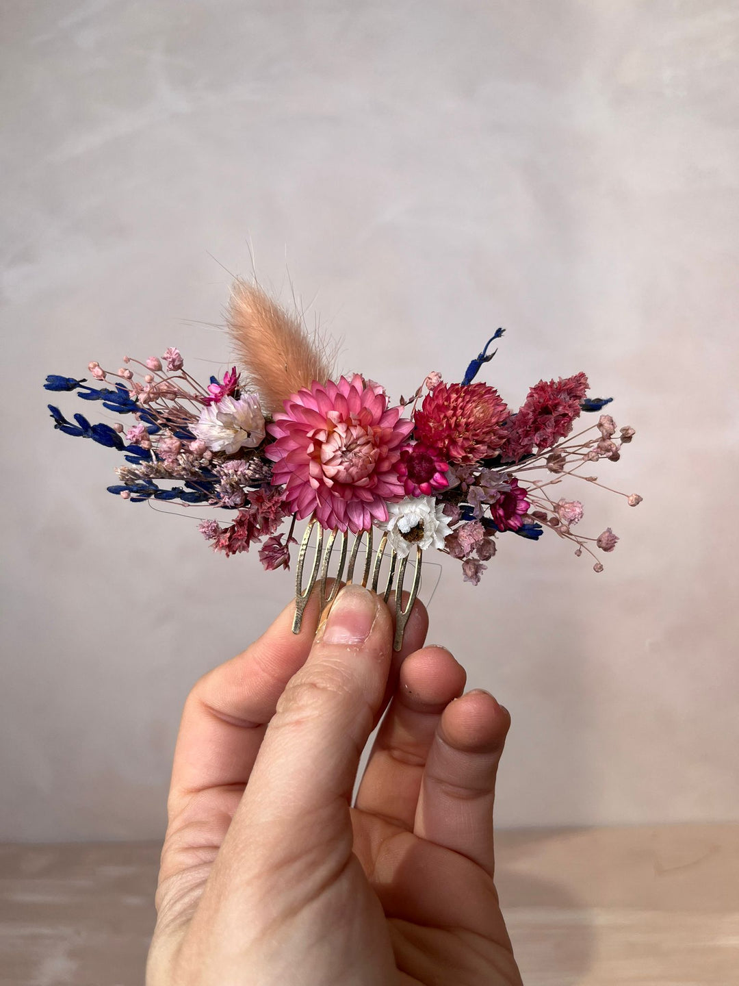A hand holding a small dried floral hair comb featuring pink and red flowers, blue accents, and a tan feather against a neutral beige background.