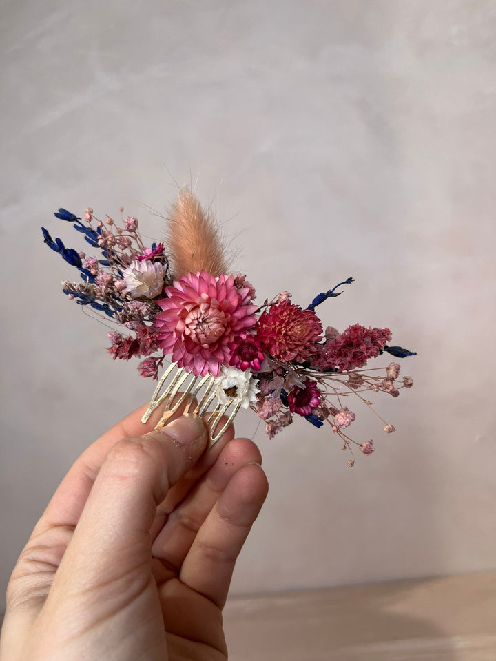 A hand holding a dried flower hair comb accessory with pink and red flowers, blue accents, and a beige feather against a neutral beige background.