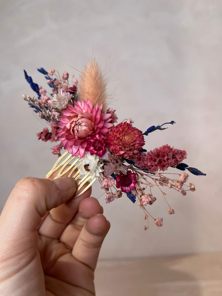 A hand holding a dried flower hair comb featuring pink and red flowers, a beige pampas grass stem, and small blue accents against a neutral beige background.