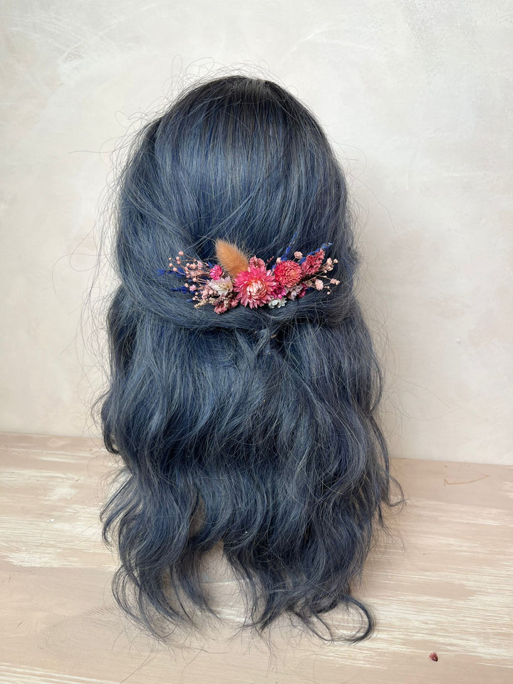 A long, wavy dark blue-gray wig displayed on a mannequin head, styled with a small floral hair accessory featuring pink, red, and brown elements, set against a neutral beige background.