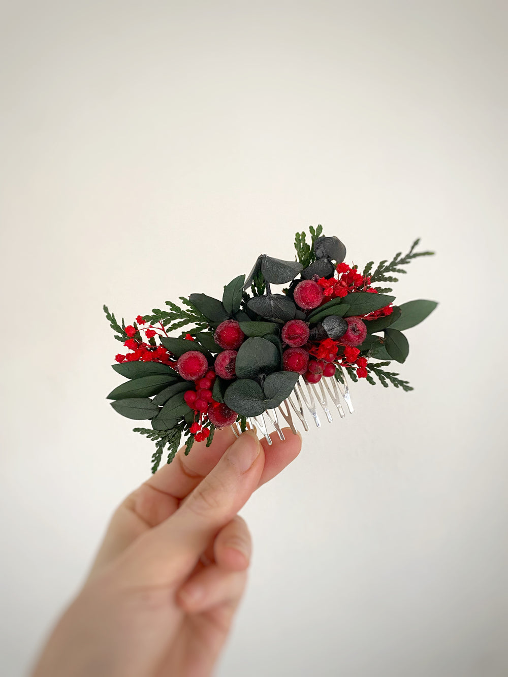 A hand holding a small decorative hair comb featuring green leaves, red berries, and dark gray accents against a plain light background.