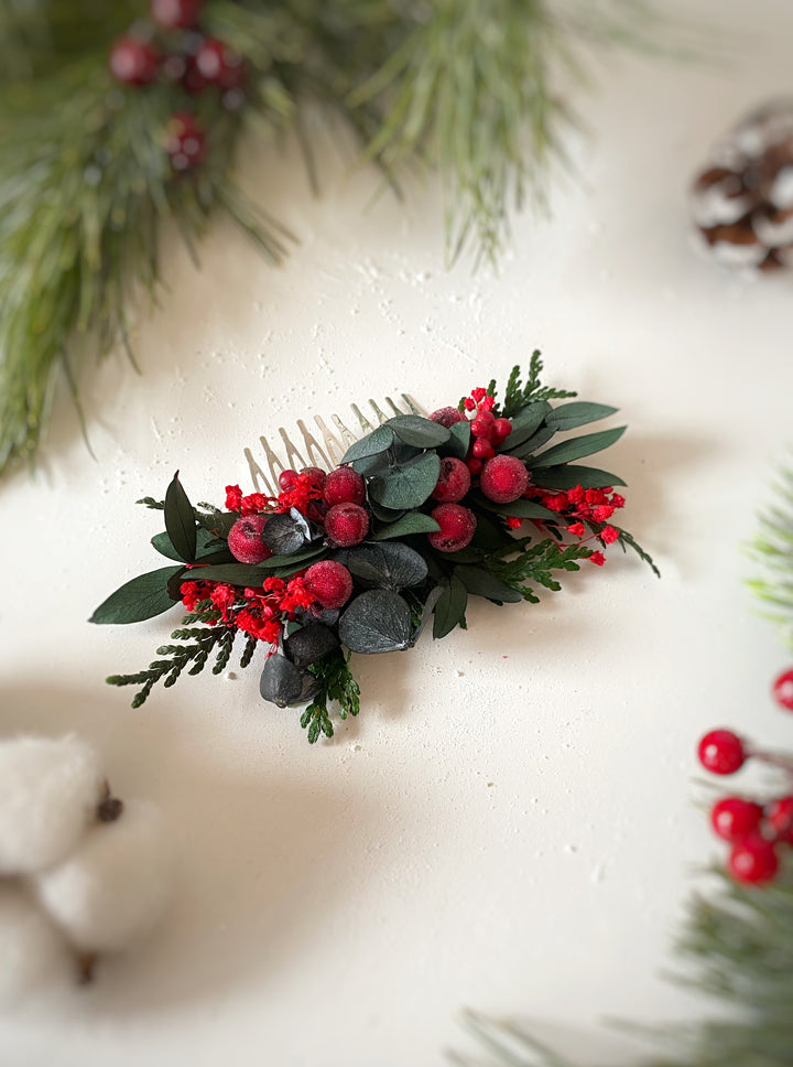 A small Christmas hair comb accessory with green eucalyptus leaves, red berries, and evergreen branches, displayed on a textured off-white surface with festive blurred background details.