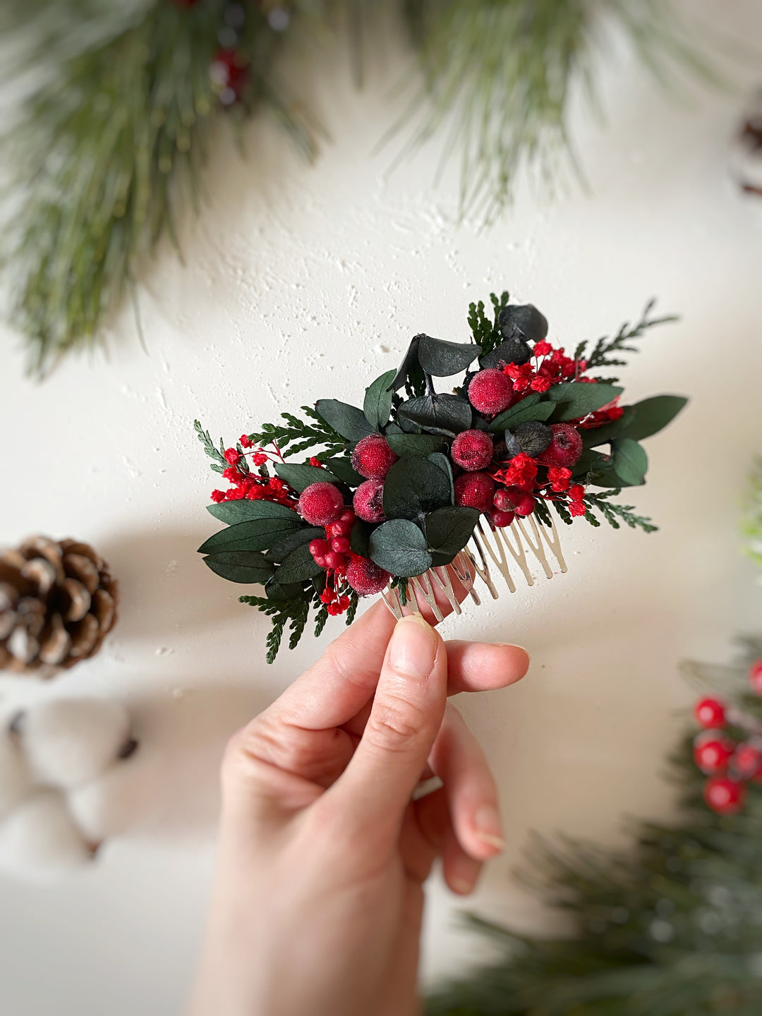 A hand holds a small decorative hair comb featuring green eucalyptus leaves and red berries, against a soft cream background with blurred holiday greenery.