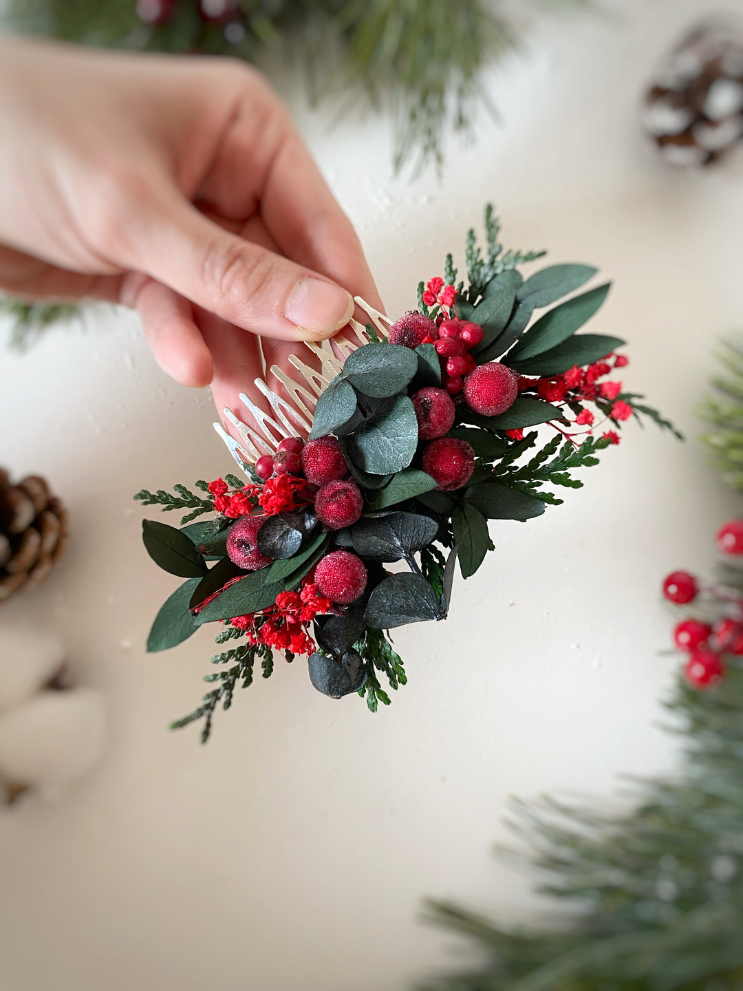 A hand holds a white hair comb decorated with green eucalyptus leaves and red berries, against a soft, light background with holiday greenery.