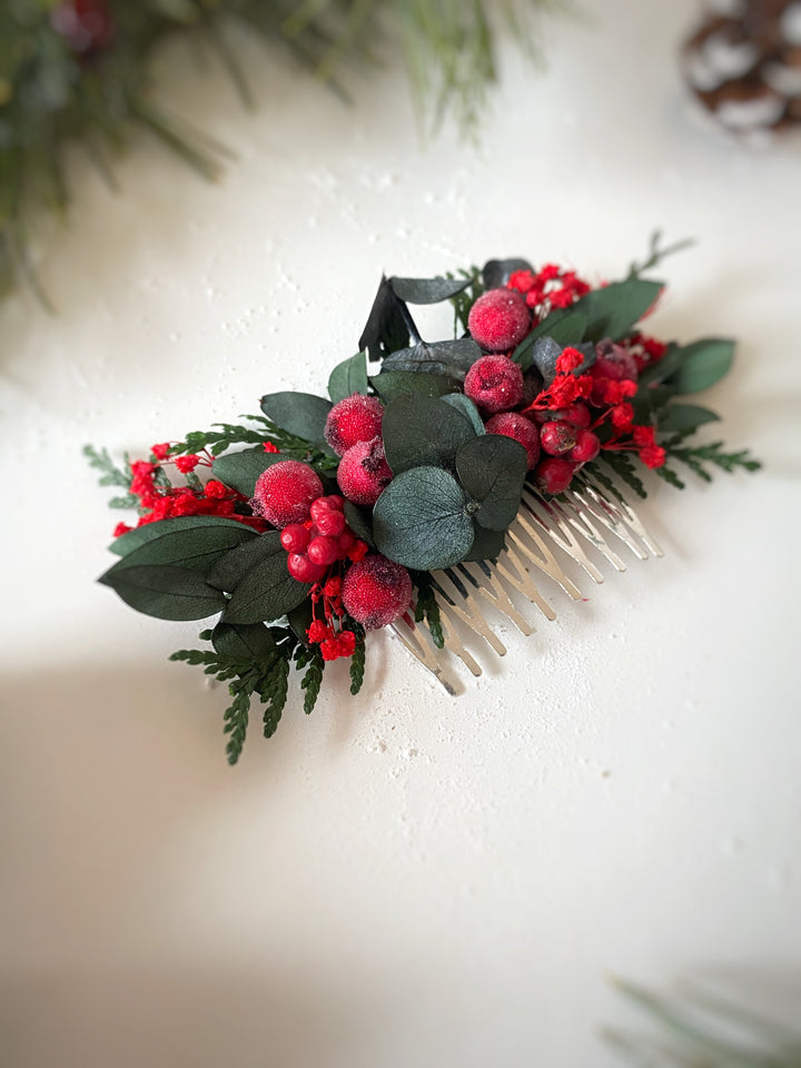 A decorative hair comb featuring a festive holiday floral design with green eucalyptus leaves, red berries, and frosted pink berries, shown on a textured off-white background.