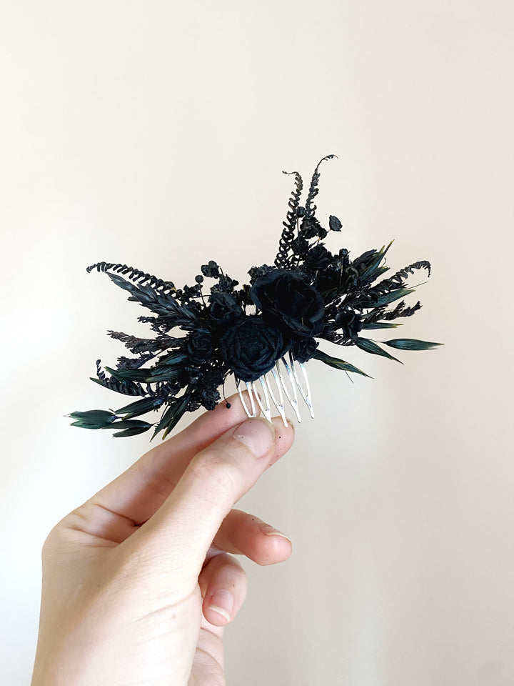 A hand holding a black floral hair comb accessory with dried ferns, small black flowers, and silver-toned metal teeth against a plain light beige background.
