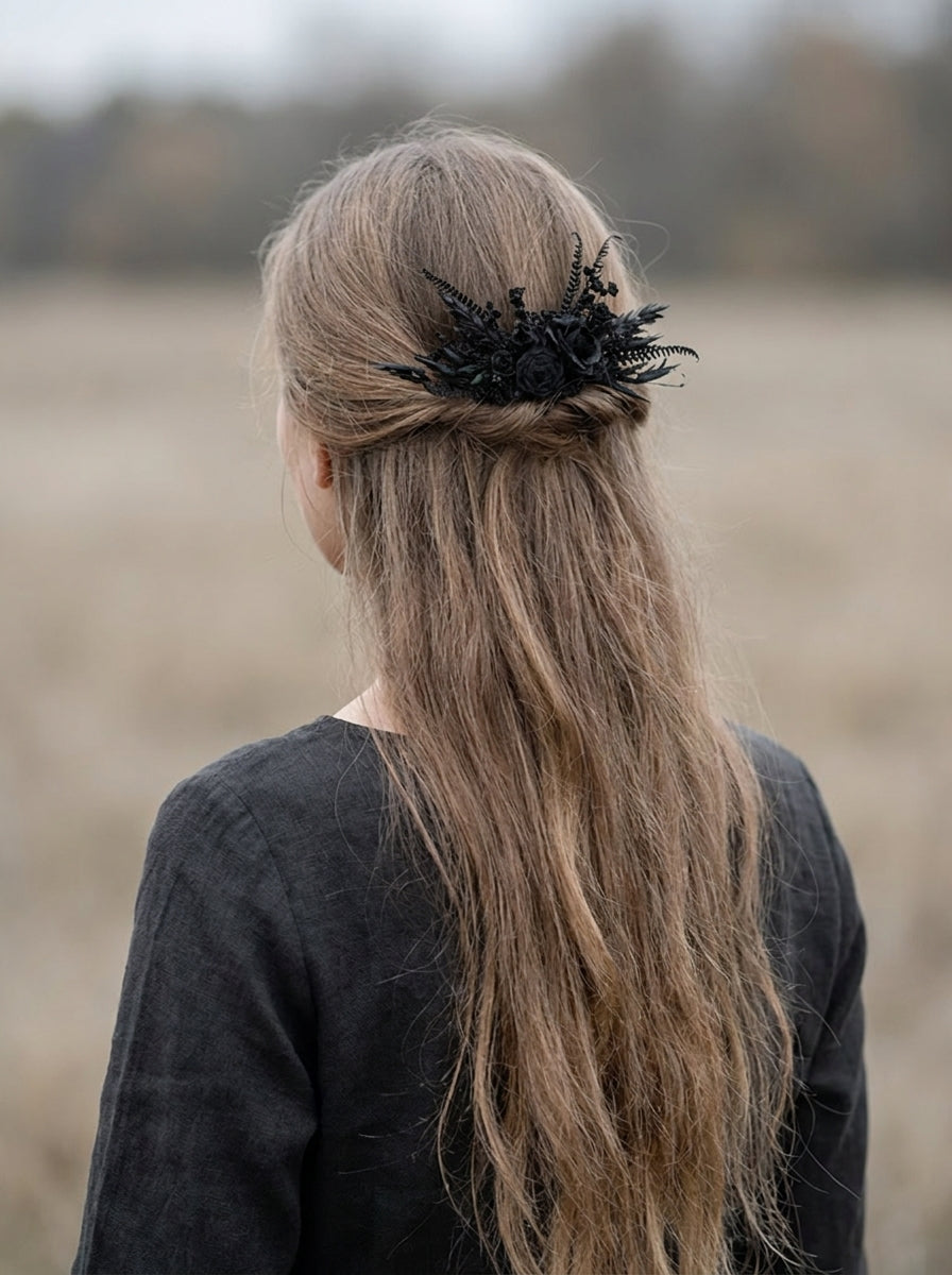 A woman viewed from behind wearing a long, light brown half-up hairstyle with a black feathered hair accessory, set against a blurred outdoor field background.