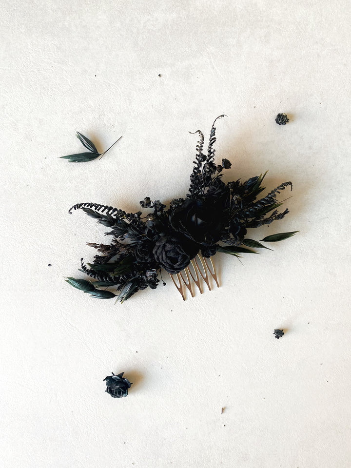 A black floral hair comb featuring dark roses, ferns, and leaves, displayed on a textured off-white background with scattered dried botanical elements.