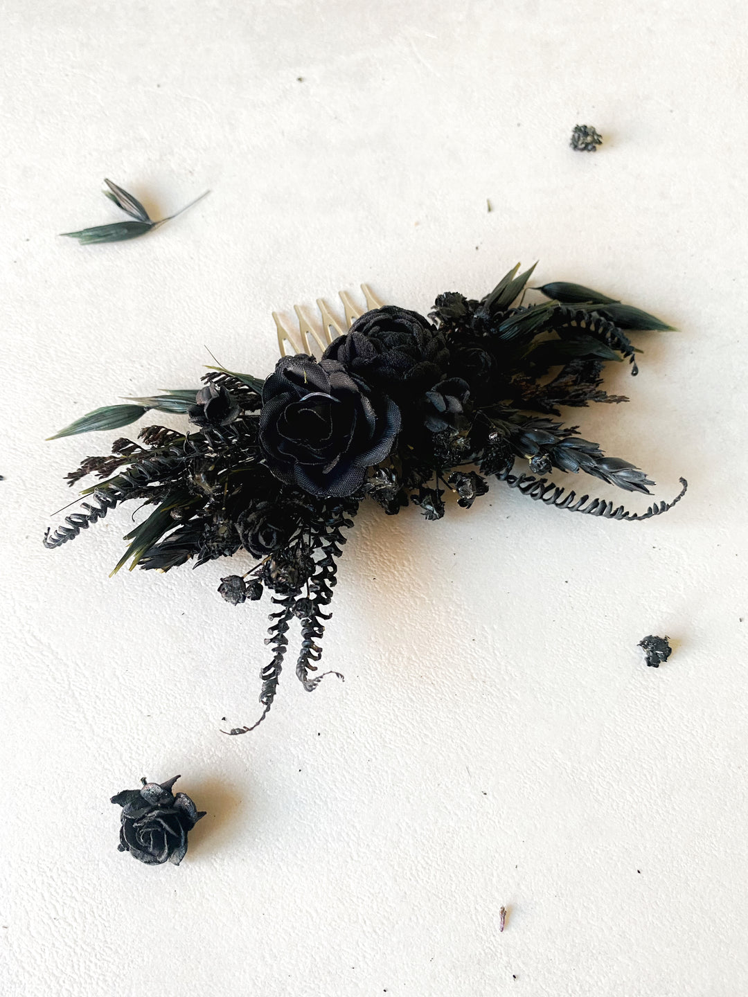A black floral hairpiece with dried ferns and a cream comb, displayed on a textured off-white background with scattered dried flower petals.