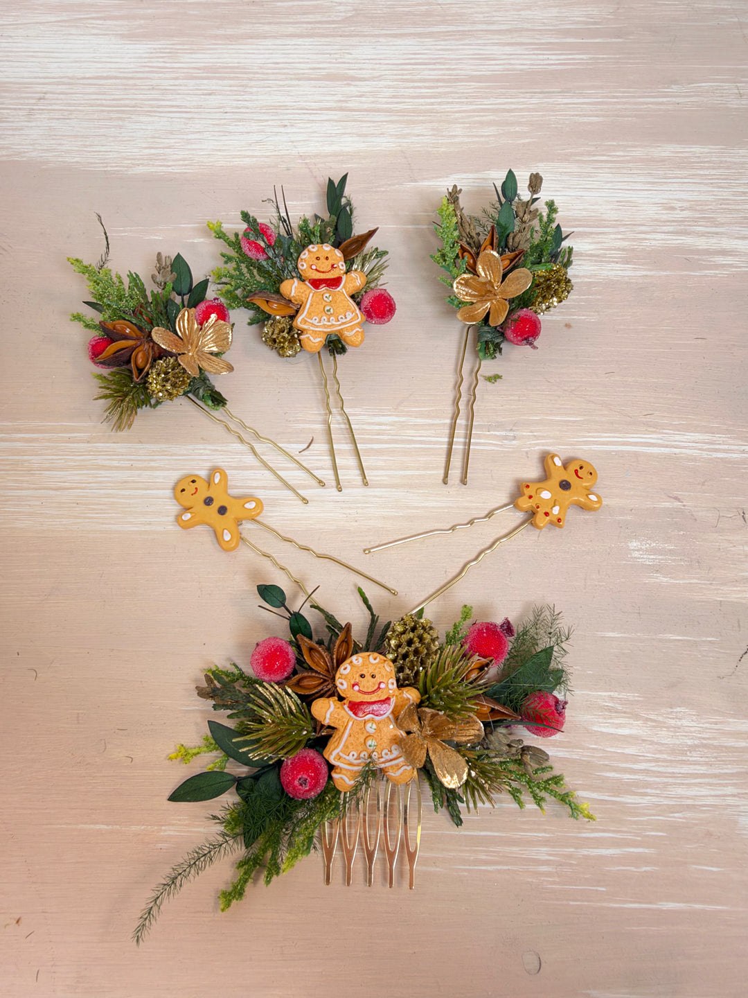 Three gingerbread-themed hair pins arranged on a light wood background. The set includes one large comb with a central gingerbread figure and surrounding greenery and berries, plus two smaller pins with single gingerbread figures attached to thin sticks.