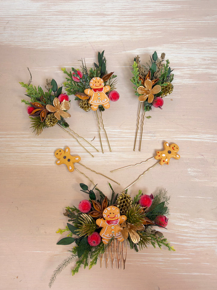 A set of Christmas-themed hair accessories arranged on a light wood background. The collection includes six  hair  pins, and one large hair comb, all decorated with green foliage, pinecones, red berries, and gingerbread men figures.
