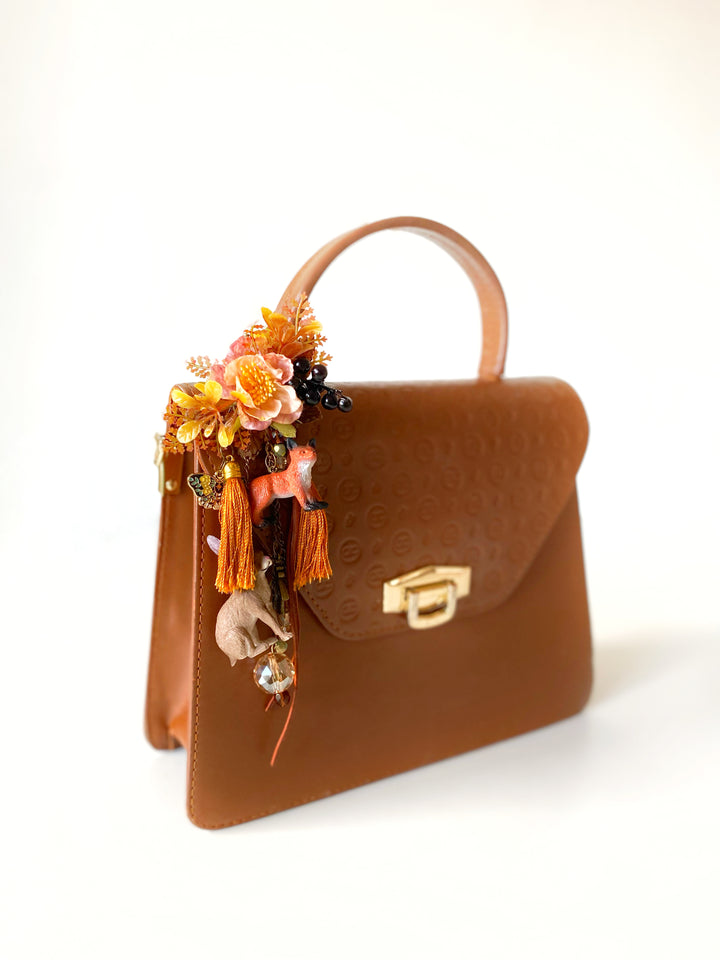 A brown leather top-handle handbag with a gold clasp, decorated on the side with a floral and tassel accessory featuring orange, yellow, and pink tones, set against a plain white background.