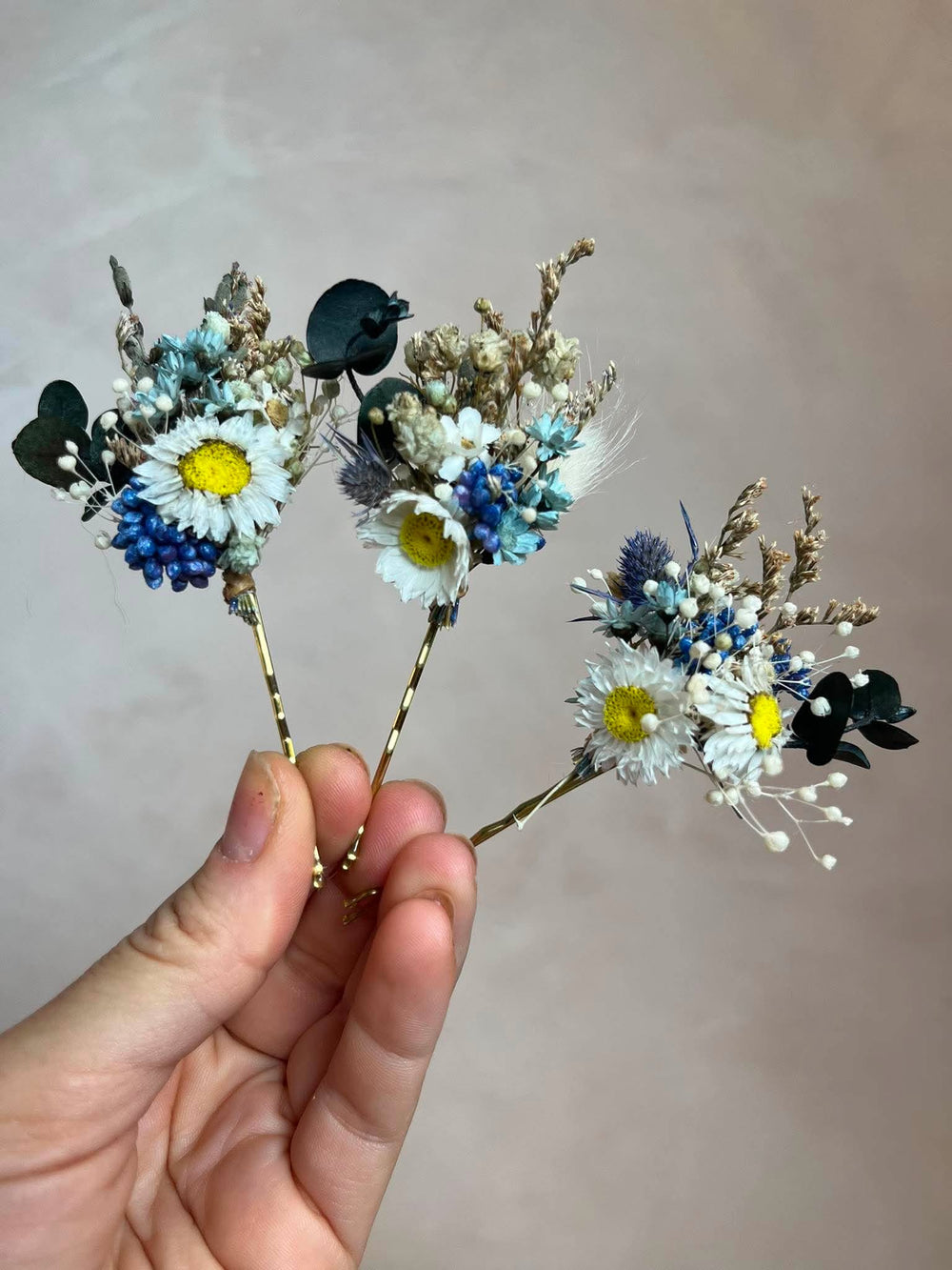 A hand holding three small dried flower hair pins with white daisies, blue grasses, and eucalyptus on gold-toned wires against a soft beige background.