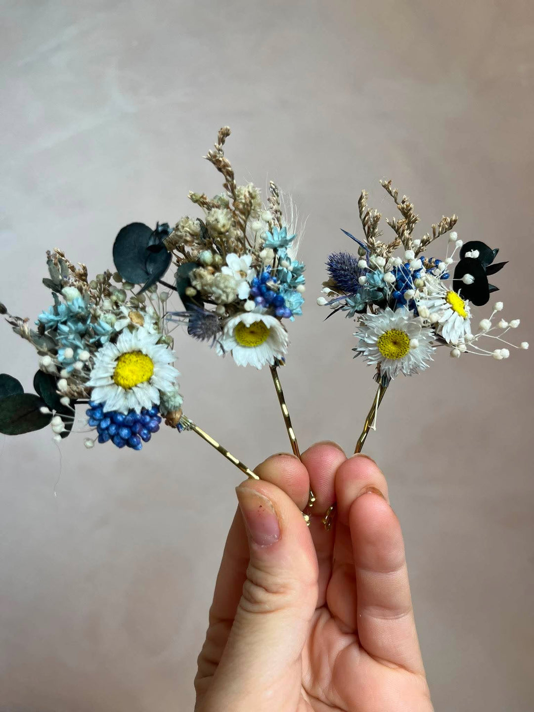 A hand holding three small dried floral hair pins with white daisies, blue blossoms, eucalyptus leaves, and tiny black flowers against a plain beige background.