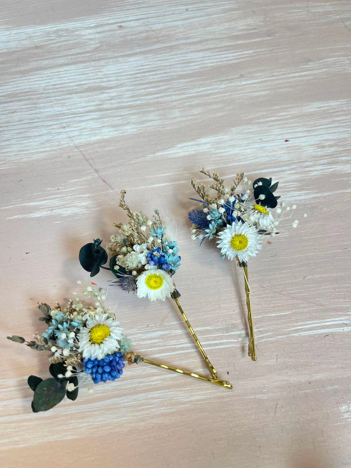 Three small dried flower hair pins with white daisy-like flowers, blue accents, and gold-tone sticks, arranged on a pale wood surface.