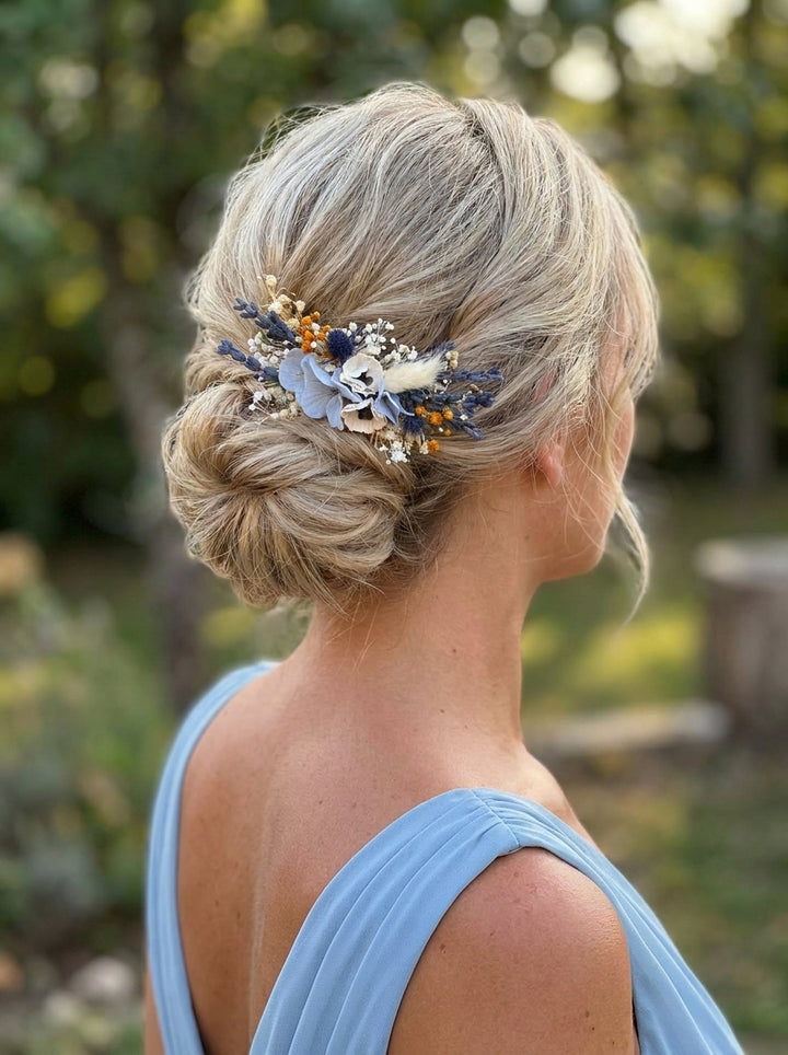 A blonde woman shown from the back wearing a light blue dress, with a decorative floral hair accessory tucked into her updo outdoors.