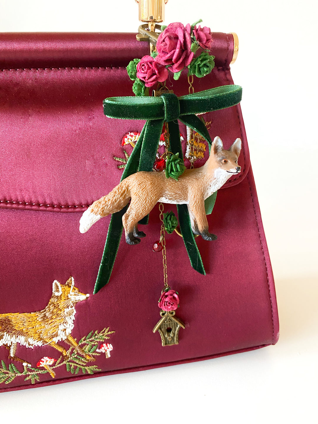 A close-up of a burgundy satin handbag with embroidered fox motifs and a decorative hanging charm featuring a small fox figurine, green ribbon, pink roses and a birdhouse.