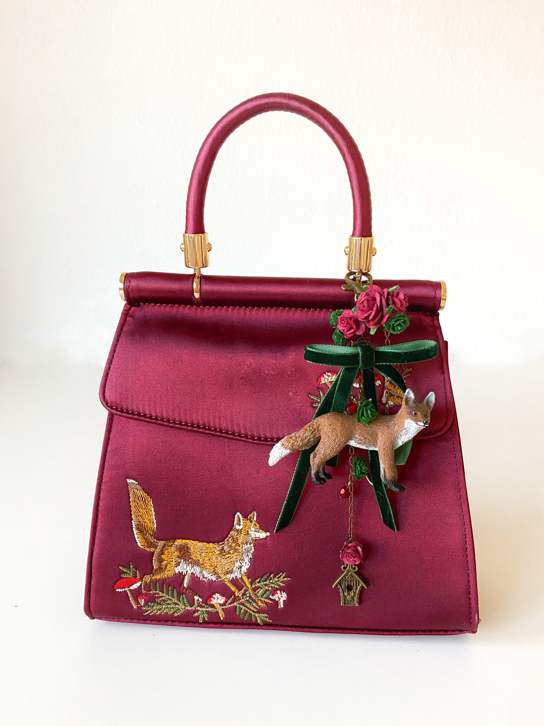 A burgundy satin handbag with gold-tone hardware, featuring embroidered fox and floral motifs on the front flap, a green ribbon bow, and a hanging fox charm.