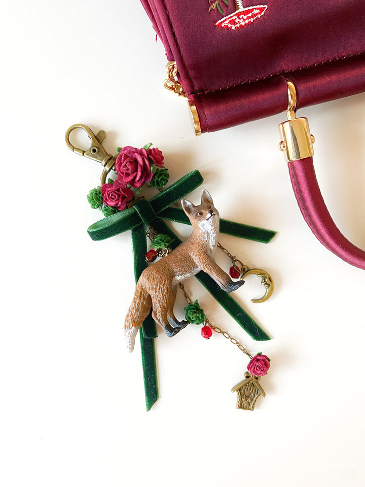 A handbag charm featuring a small fox figurine standing on a green tree with pink roses, gold-tone hardware, and a small birdhouse charm, shown next to a burgundy handbag on a white background.