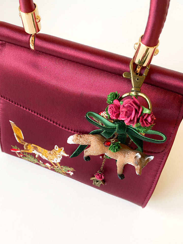 A close-up of a burgundy satin handbag with gold-tone hardware, featuring an embroidered fox motif on the front pocket and a decorative hanging charm with pink roses, a green ribbon, and a small fox figure.