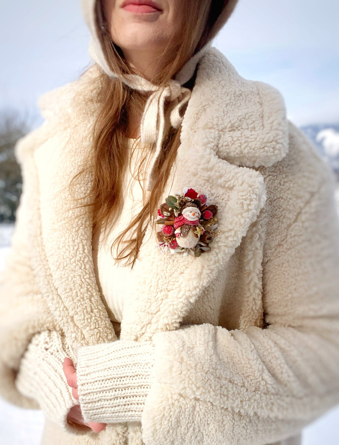 A close-up of a person wearing a cream sherpa fleece coat over a cream knit top, with a small holiday snowman brooch pinned to the chest, set against a snowy outdoor background.