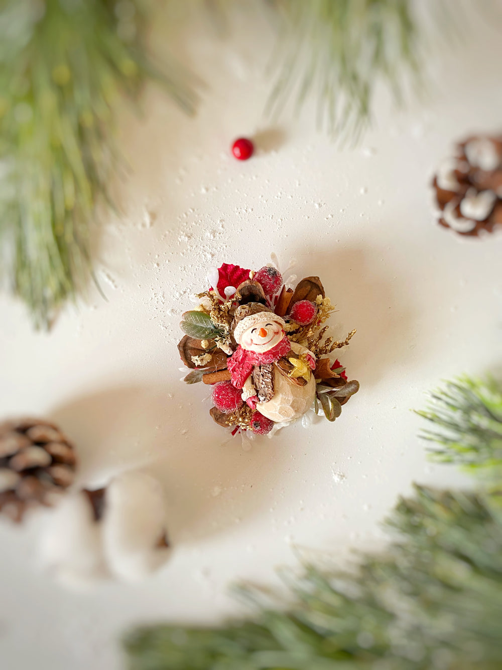 A small decorative snowman ornament nestled in red berries, brown pinecones, and green foliage, arranged on a white surface with blurred evergreen branches and pinecones around it.