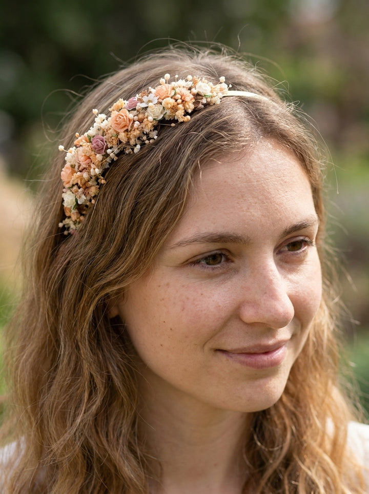 A woman with long wavy light brown hair wearing a delicate floral headband with peach, cream, and pink blossoms, photographed outdoors with a soft blurred green background.