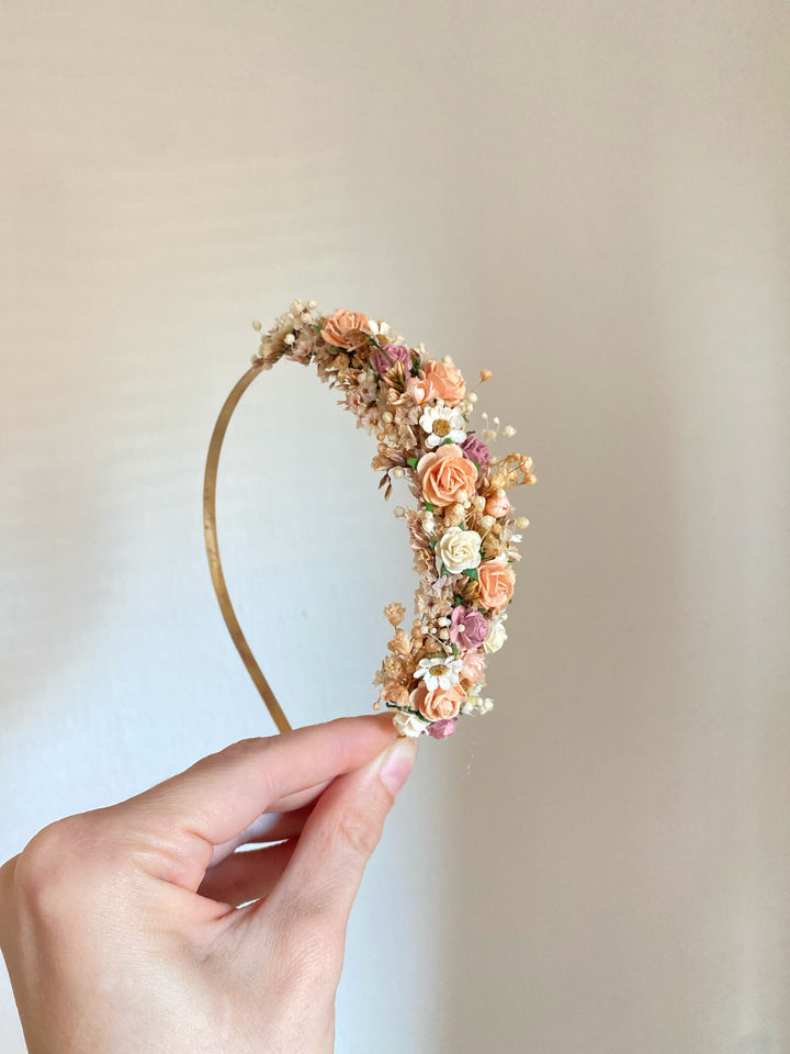 A hand holding a gold metal headband decorated with peach, pink, and cream flowers against a plain beige background.