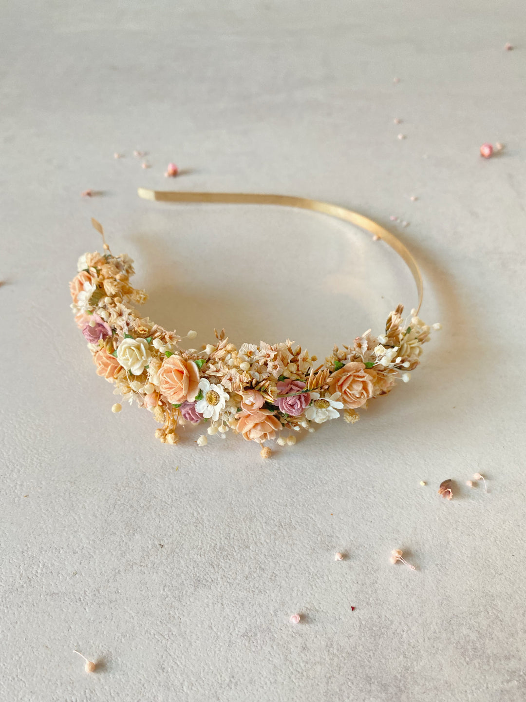 A gold metal headband decorated with peach roses, small white flowers, and green foliage, displayed on a textured light gray surface.