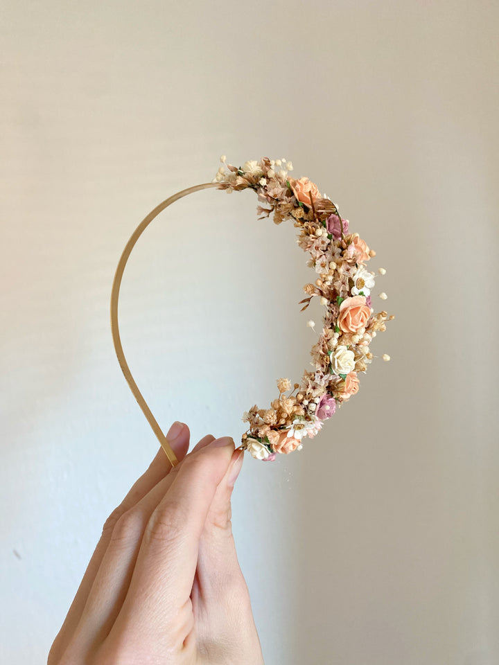 A hand holding a gold-tone crescent moon headband decorated with peach, pink, and cream flowers against a plain beige wall.