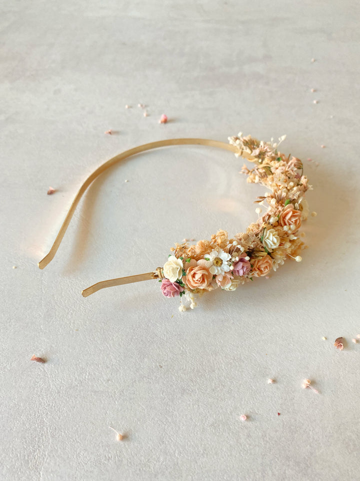 A delicate floral headband with a curved gold-toned band, decorated with small or pressed flowers in soft peach, cream, pink, and white, photographed on a light neutral surface.