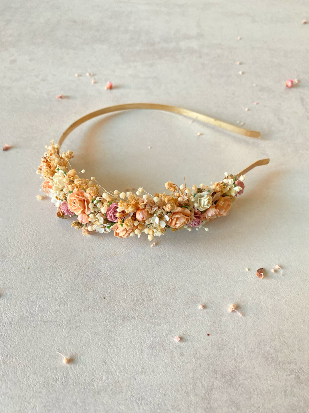 A gold metal headband decorated with peach roses, baby's breath, and small pink flowers, displayed on a textured light gray surface.
