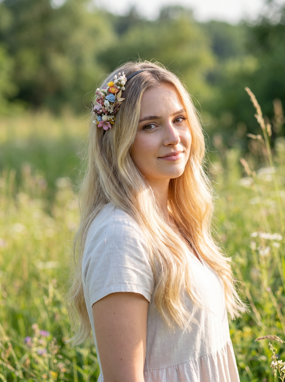 A young blonde woman wearing a delicate pastel flower crown and a light-colored short-sleeve dress, standing in a sunlit grassy field.