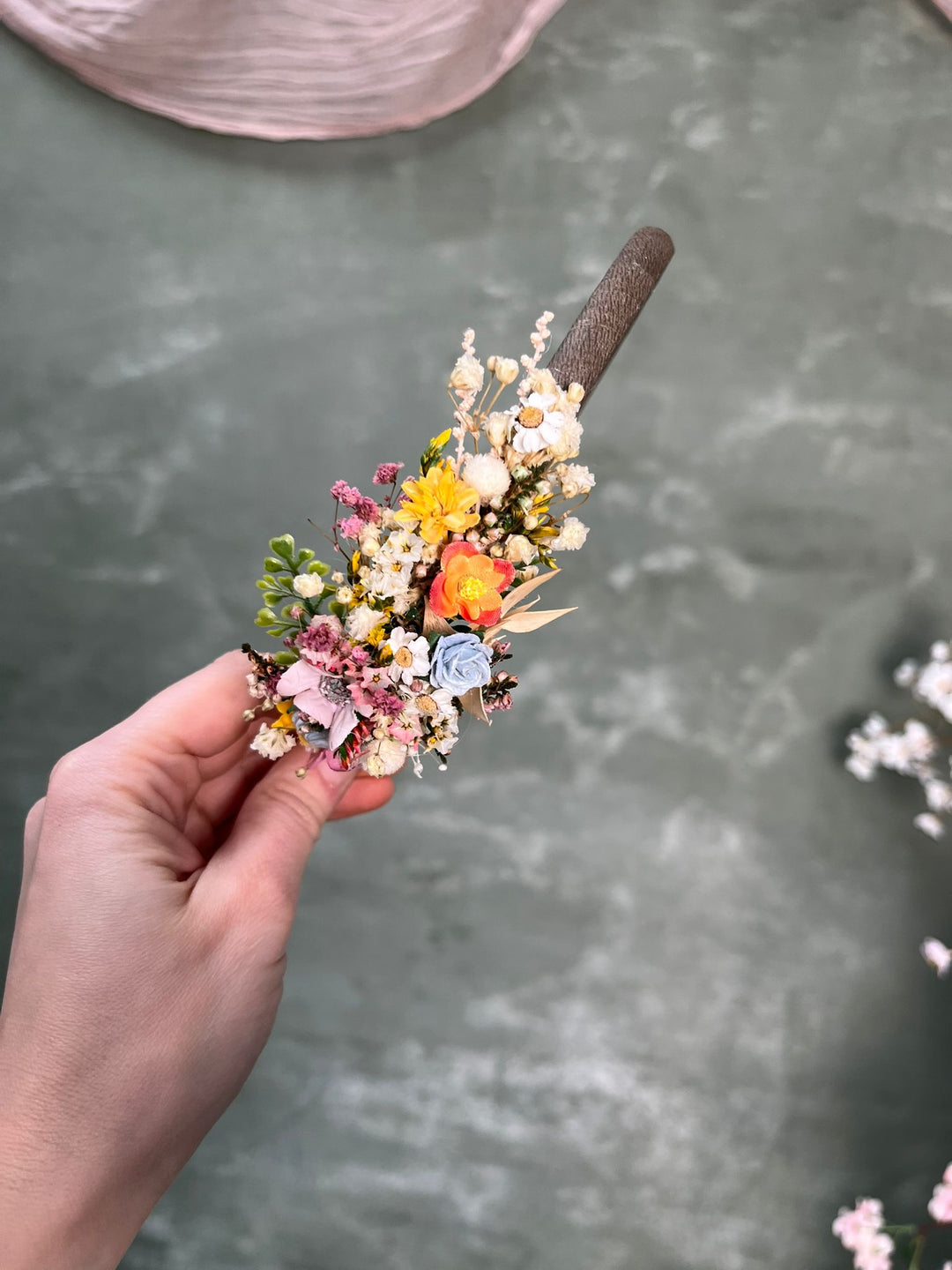 A hand holding a small flower headband accessory with a brown textured band, featuring delicate pastel and bright flowers including yellow, pink, orange, and blue on a gray textured background.