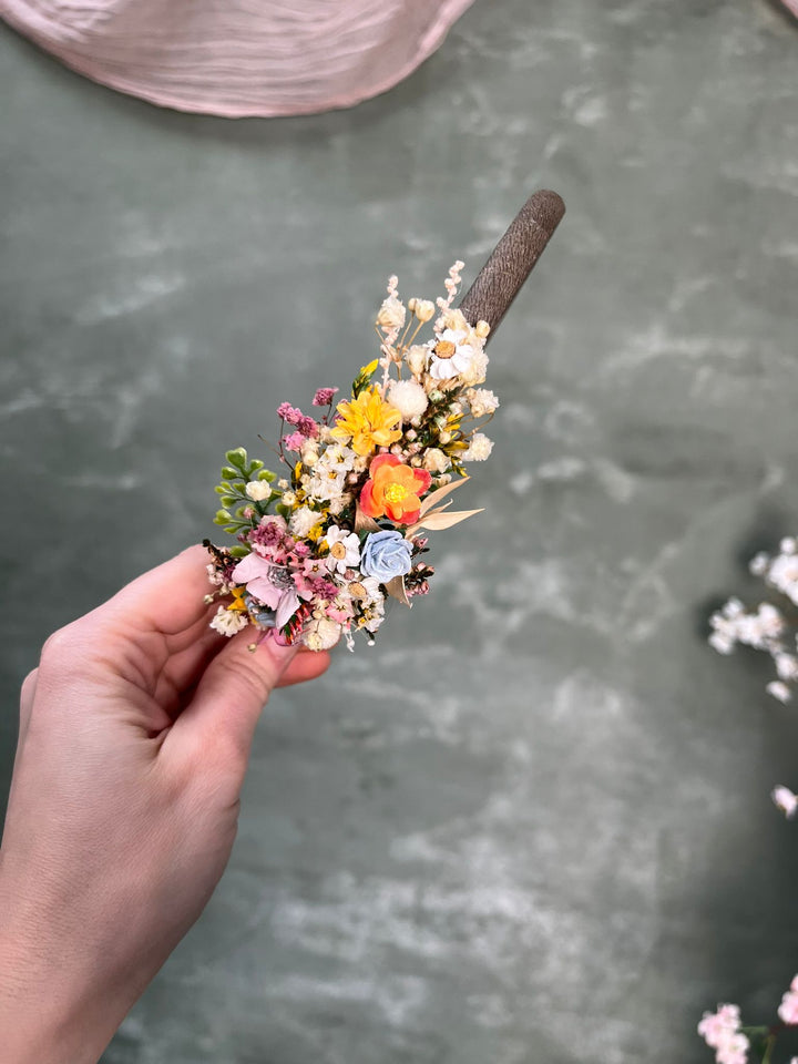 A hand holding a small flower headband accessory with a brown textured band, featuring delicate pastel and bright flowers including yellow, pink, orange, and blue on a gray textured background.