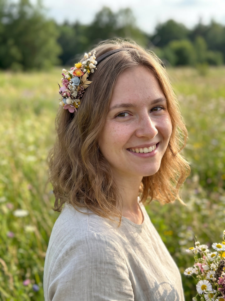 A smiling woman with shoulder-length wavy light brown hair wearing a pastel floral headpiece and a light beige linen top, standing in a sunlit grassy field.