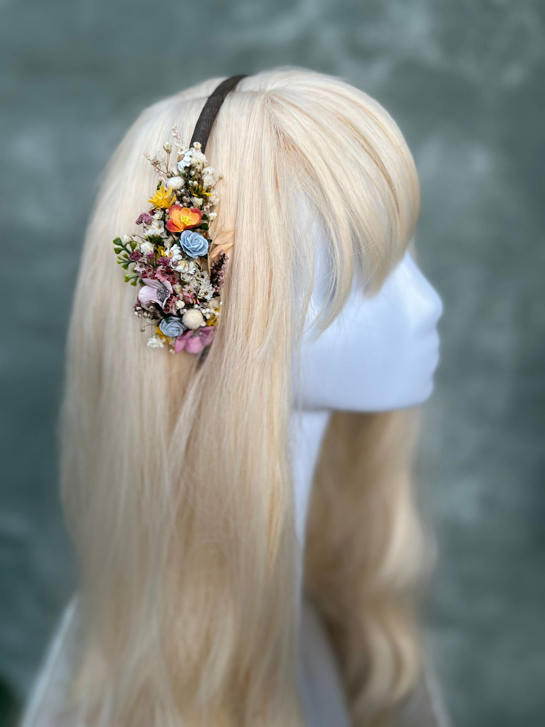A long blonde wig displayed on a white mannequin head, styled with a dark brown headband and a colorful floral hair accessory featuring flowers and small green leaves.