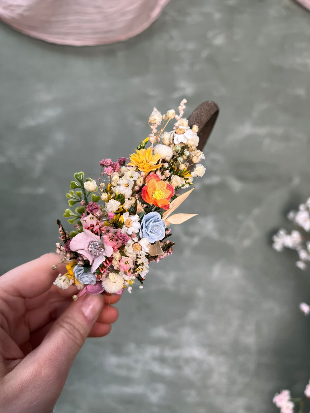 A hand holding a dark brown headband decorated with a dense cluster of small artificial flowers in pastel pink, blue, yellow, orange, and cream tones, set against a textured gray-green background.