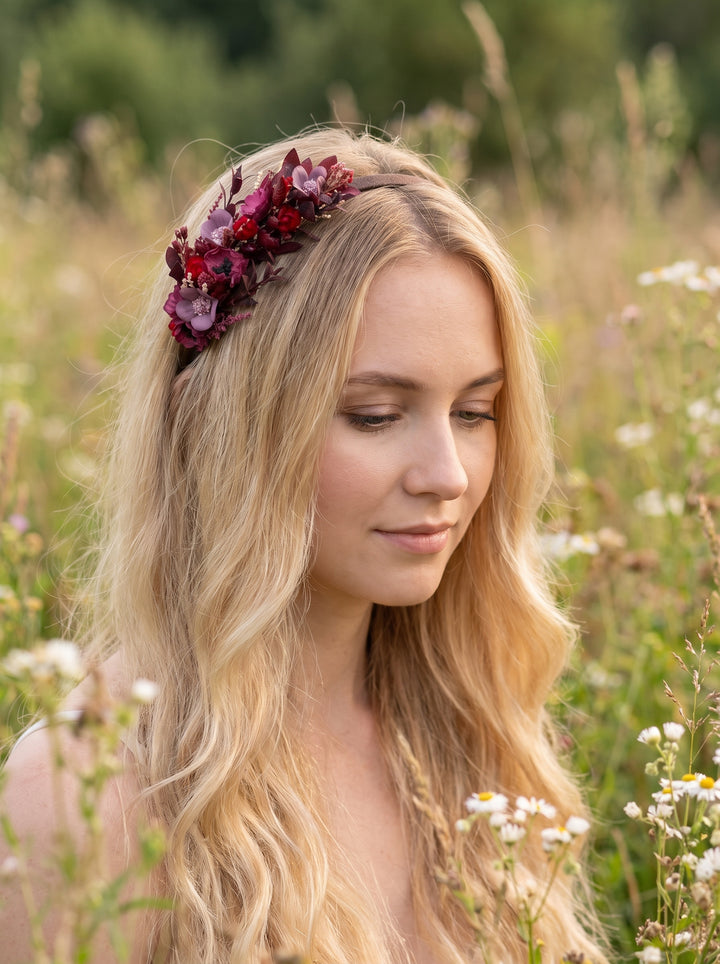A woman with long blonde hair wearing a floral crown made of dark red and purple flowers, standing in a sunlit grassy field.