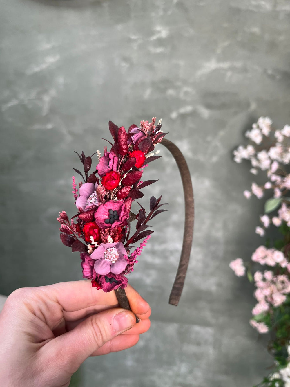 A hand holding a brown fabric headband decorated with a dried floral bouquet in shades of pink, red, and burgundy, set against a gray textured background with blurred pink flowers on the right.