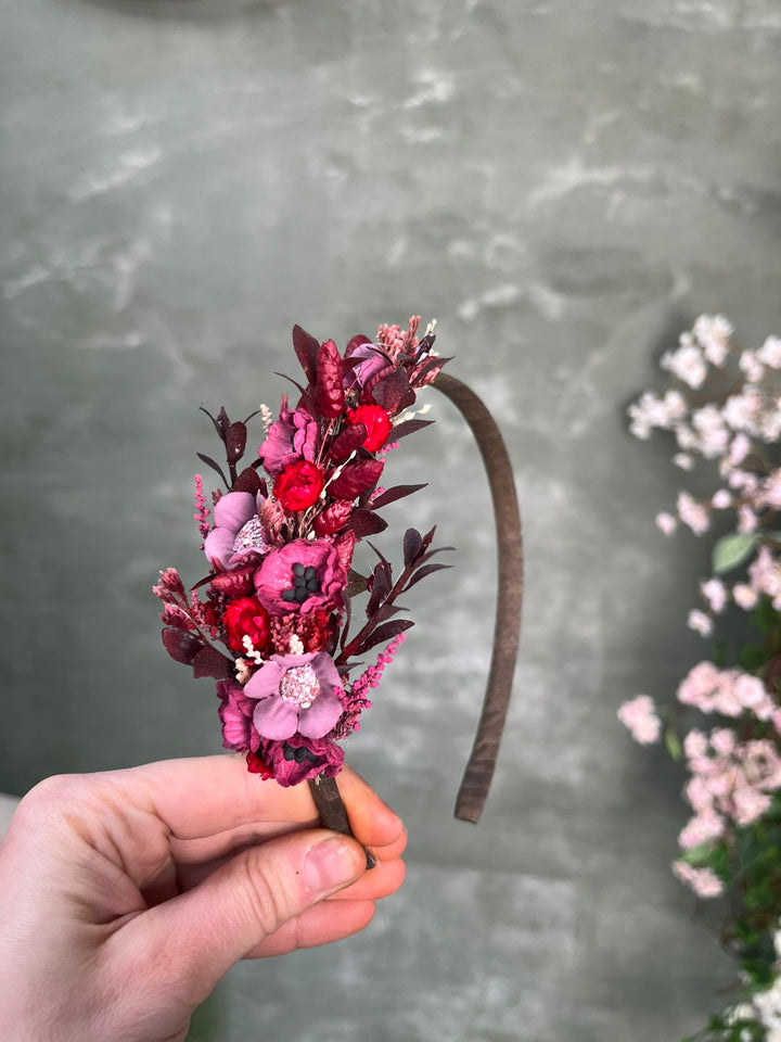 A hand holding a brown fabric headband decorated with a dried floral bouquet in shades of pink, red, and burgundy, set against a gray textured background with blurred pink flowers on the right.