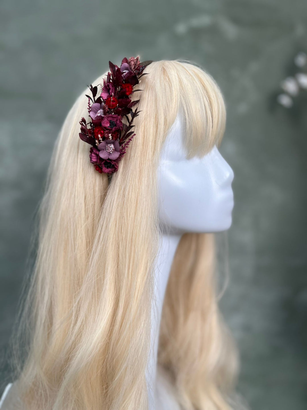 A blonde wig displayed on a white mannequin head, featuring a side profile view and a dark burgundy floral hair accessory on the left side.