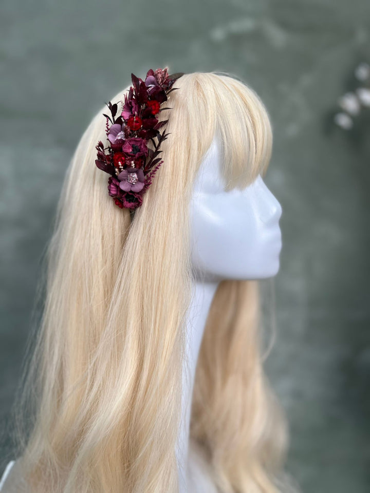 A blonde wig displayed on a white mannequin head, featuring a side profile view and a dark burgundy floral hair accessory on the left side.