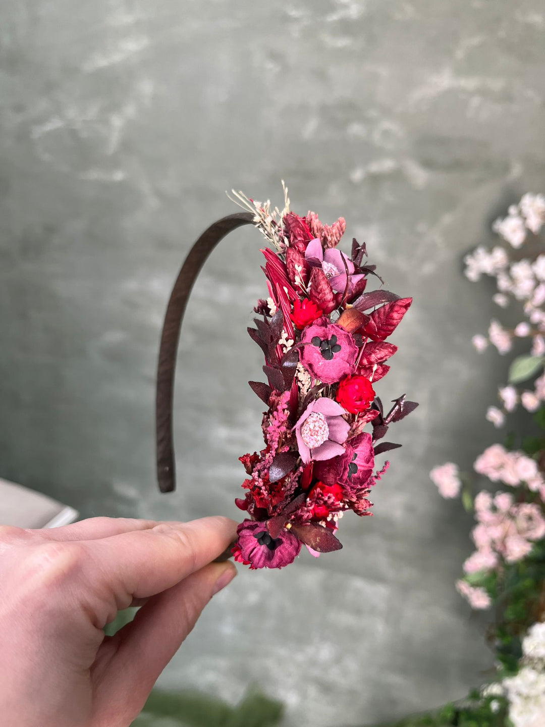 A hand holding a dark brown headband decorated with purple and red flowers and leaves, set against a gray stone wall with blurred pink flowers in the background.