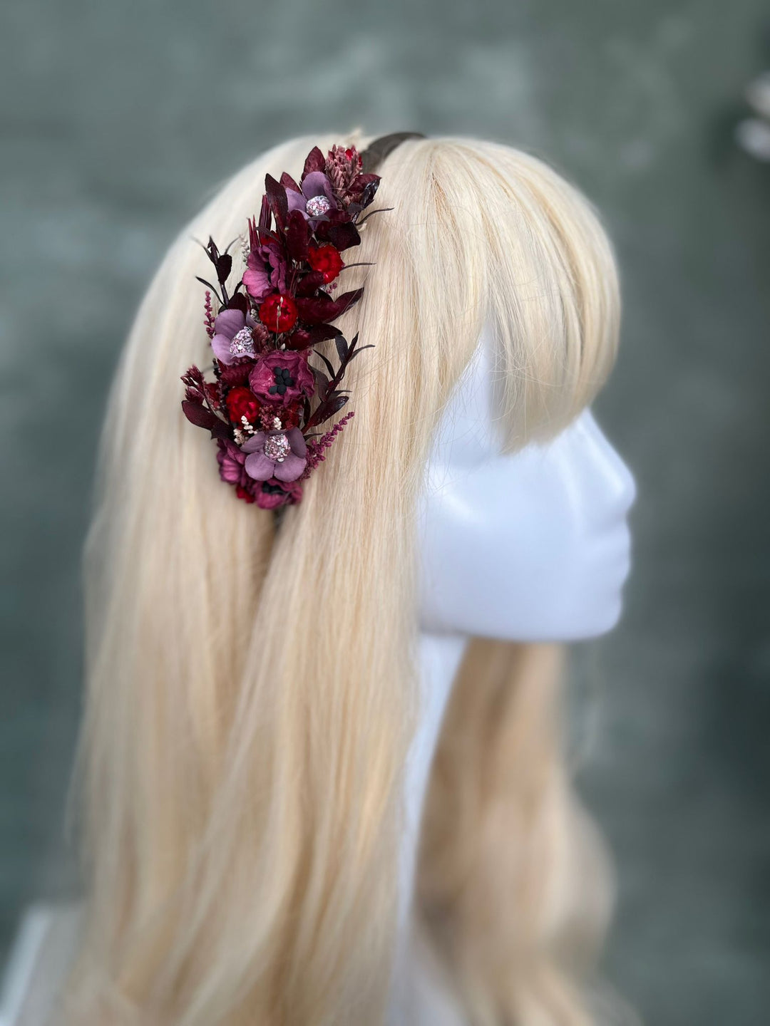 A floral hair accessory with dark burgundy, purple, and red flowers, displayed on a blonde mannequin head against a textured gray background.