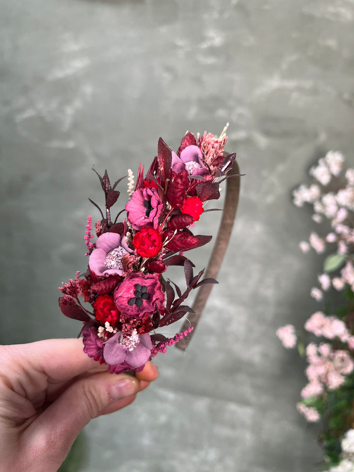 A hand holding a decorative headband adorned with flowers in shades of purple, red, and burgundy, set against a soft-focus gray background with blurred pink flowers on the right.