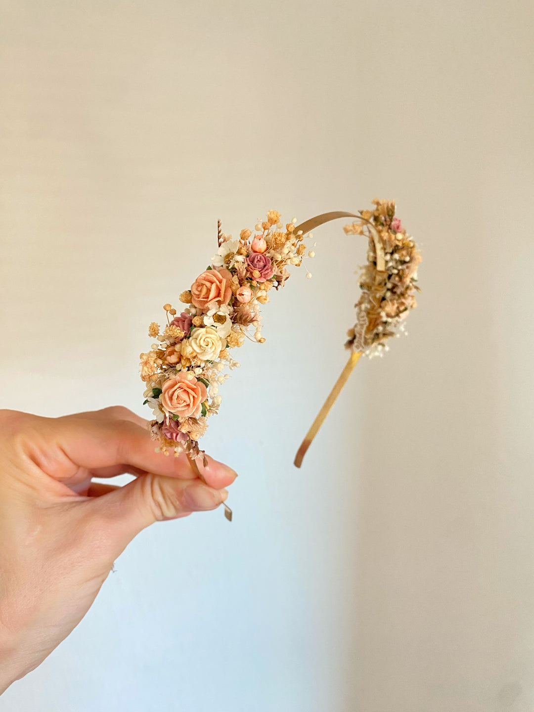 A hand holding a gold headband decorated with peach, pink, and cream flowers, against a plain light beige background.