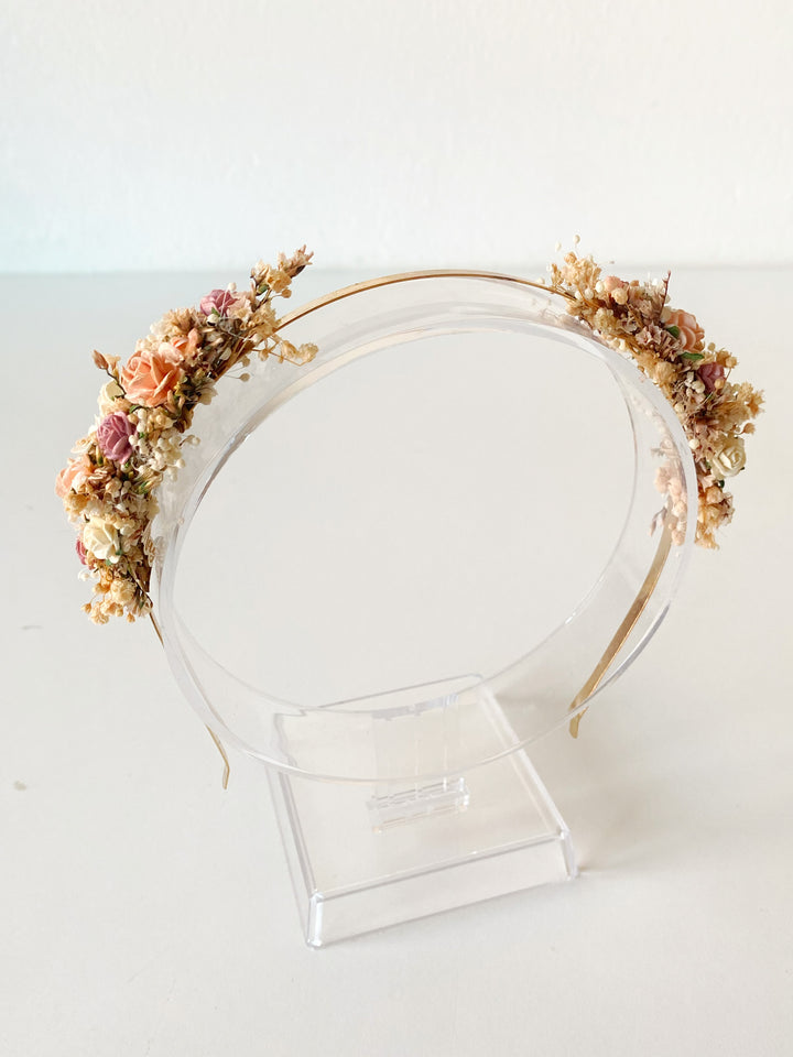 A delicate gold wire headband decorated with flowers in shades of beige, pink, and brown, displayed on a clear acrylic stand against a plain white background.