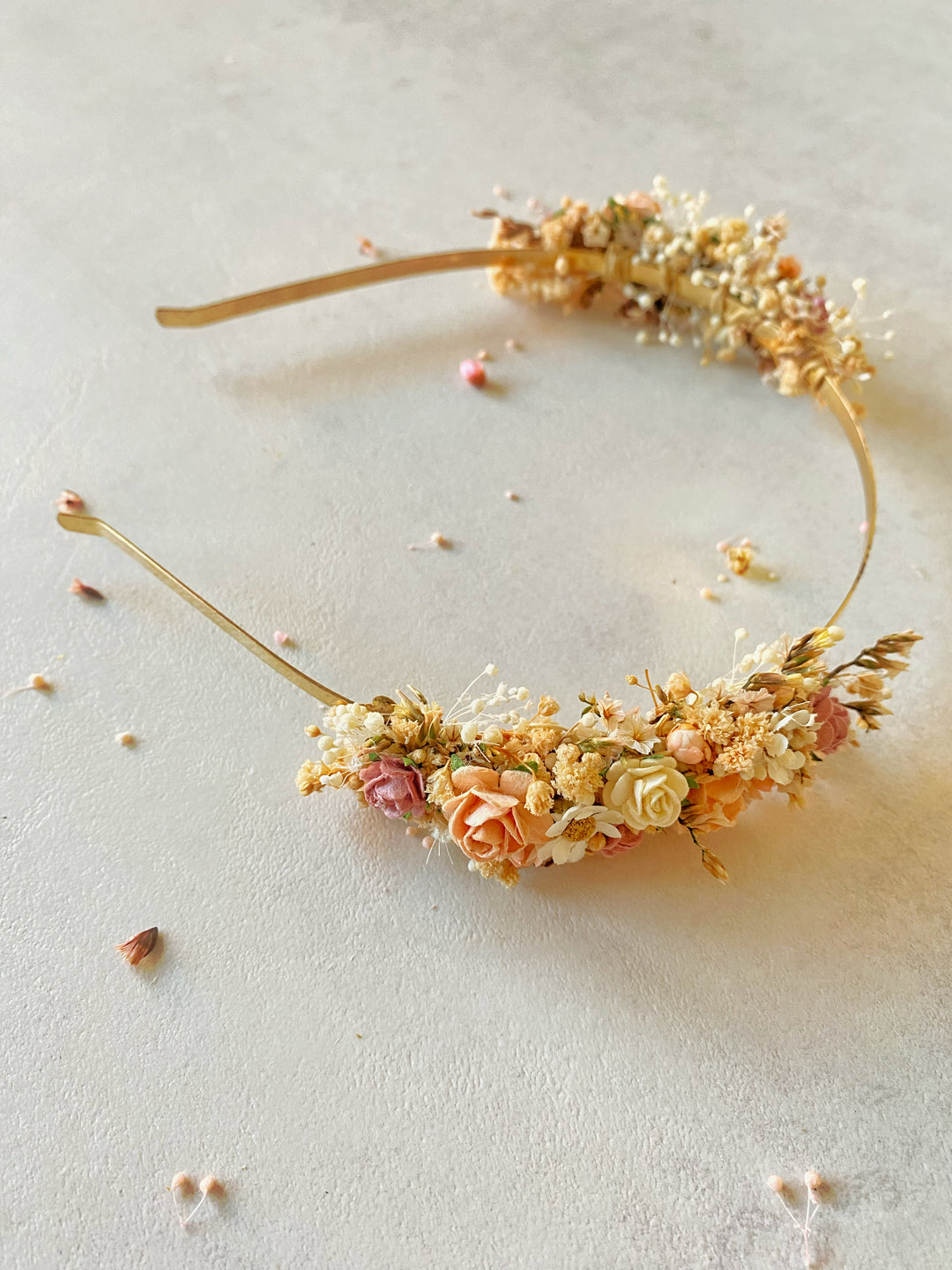 A gold metal headband decorated with dried flowers in soft peach, cream, and muted pink tones, arranged in a curved shape on a light neutral surface.